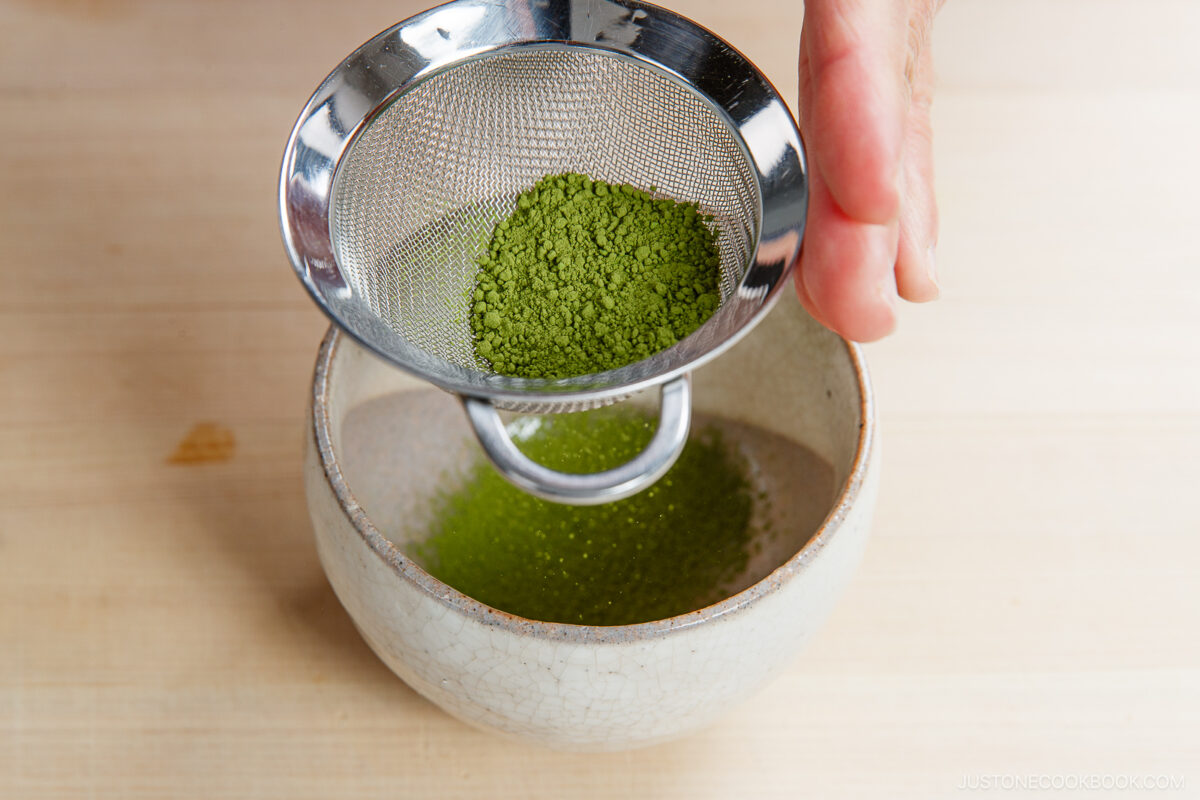 A hand holds a metal sifter over a white ceramic bowl, gently sifting vibrant green matcha powder—perfect for crafting a smooth matcha latte—onto a light wooden surface.