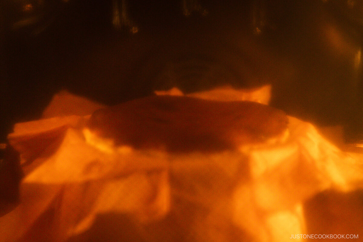 A basque cheesecake baking in an oven, photographed through the oven door with a warm, hazy glow and wrapped in parchment paper.