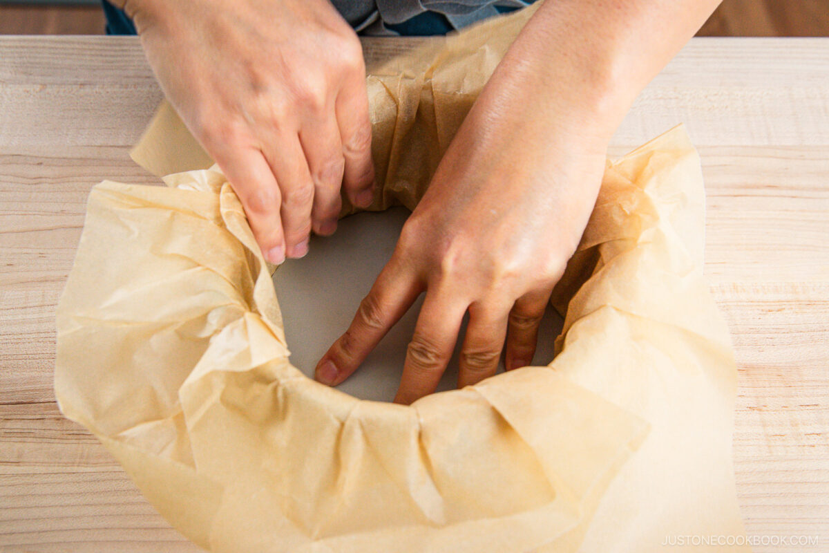Hands pressing parchment paper into a round cake pan on a wooden surface, preparing the pan for baking a Basque cheesecake.