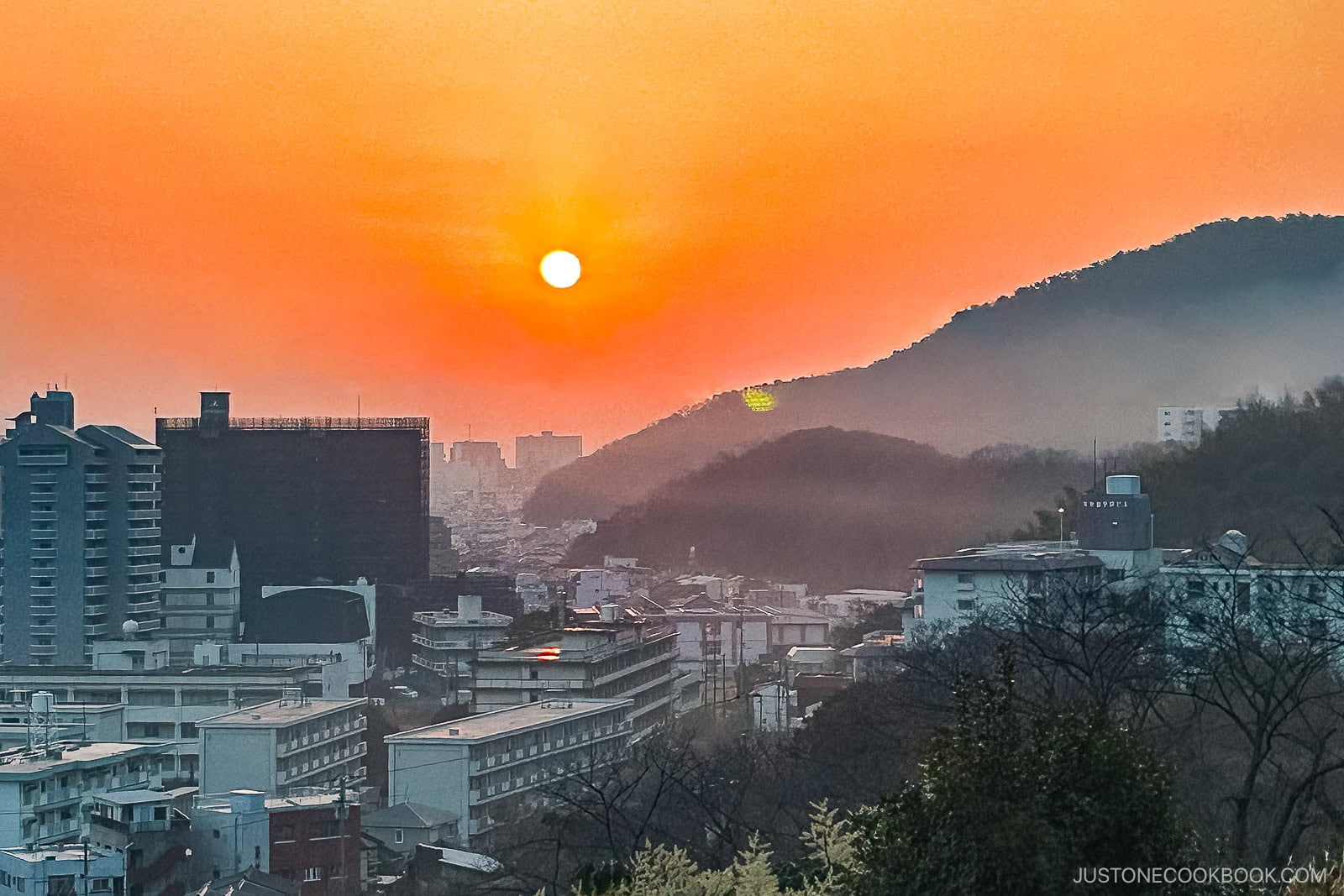 Sunset over a city with buildings in the foreground, misty hills beyond, and an orange sky evokes the serenity of Japanese New Year – Oshogatsu, as the low sun casts a warm festive glow across the tranquil landscape.