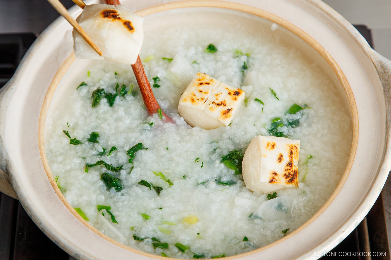 A pot of nanakusa gayu rice porridge with leafy greens and grilled white fish pieces, with chopsticks lifting one fish piece above the dish.