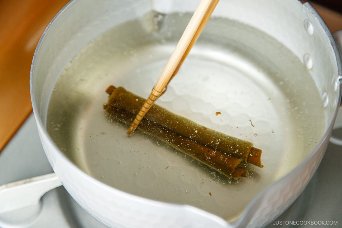 Chopsticks hold pieces of kombu (seaweed) in a pot of water on a stovetop, as part of preparing Japanese dashi broth for toshikoshi soba.