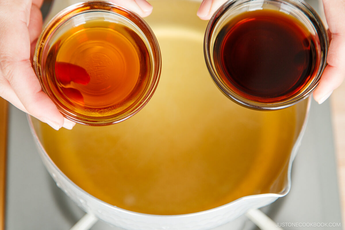 Two hands hold bowls of mirin and soy sauce above a pot, ready to pour the flavorful ingredients into toshikoshi soba simmering on the stovetop.