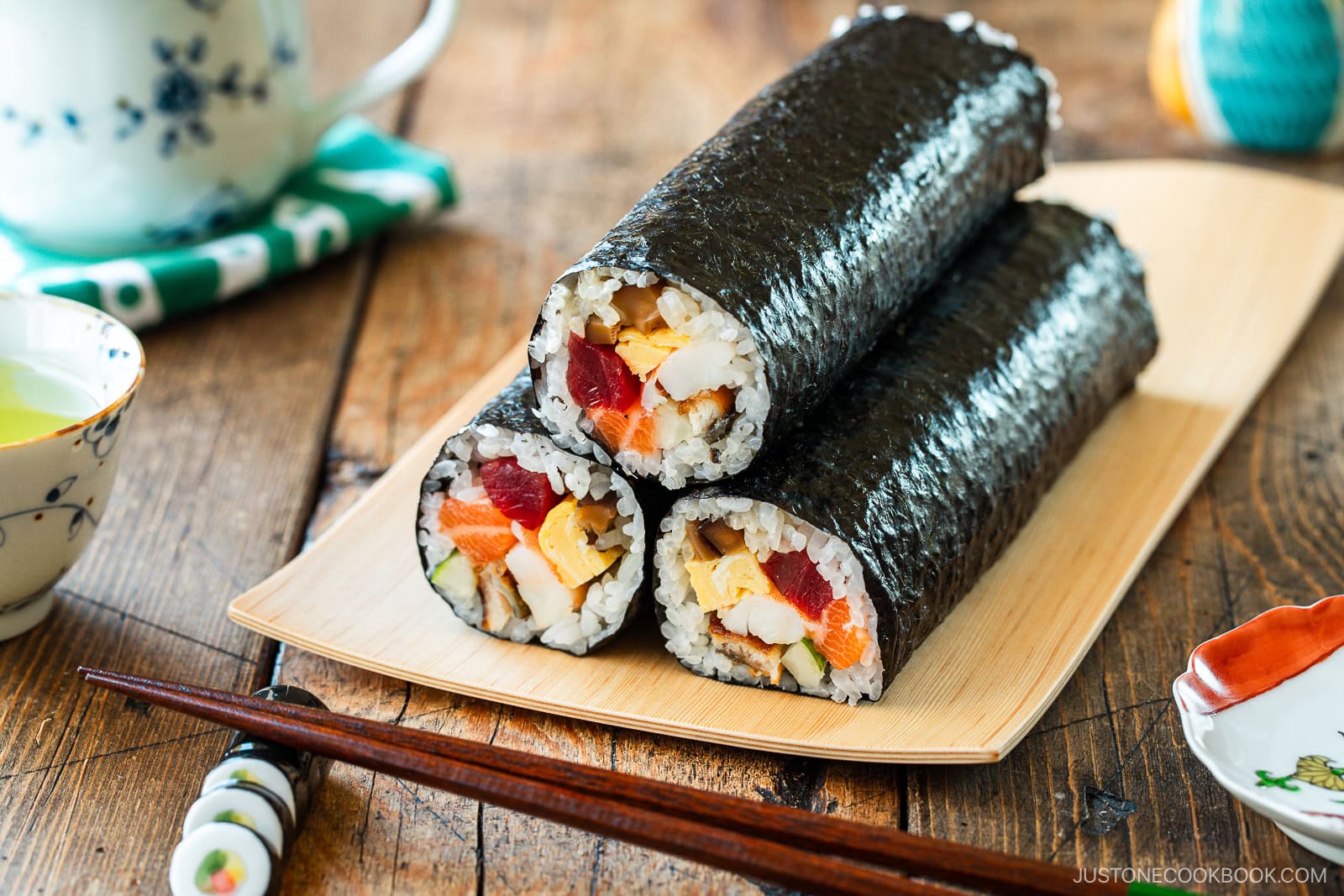 Three thick ehomaki (setsubun sushi roll) packed with colorful fish, egg, and vegetables are stacked on a wooden plate. Chopsticks, a tea cup, and small dishes complete the scene on a rustic wooden table.