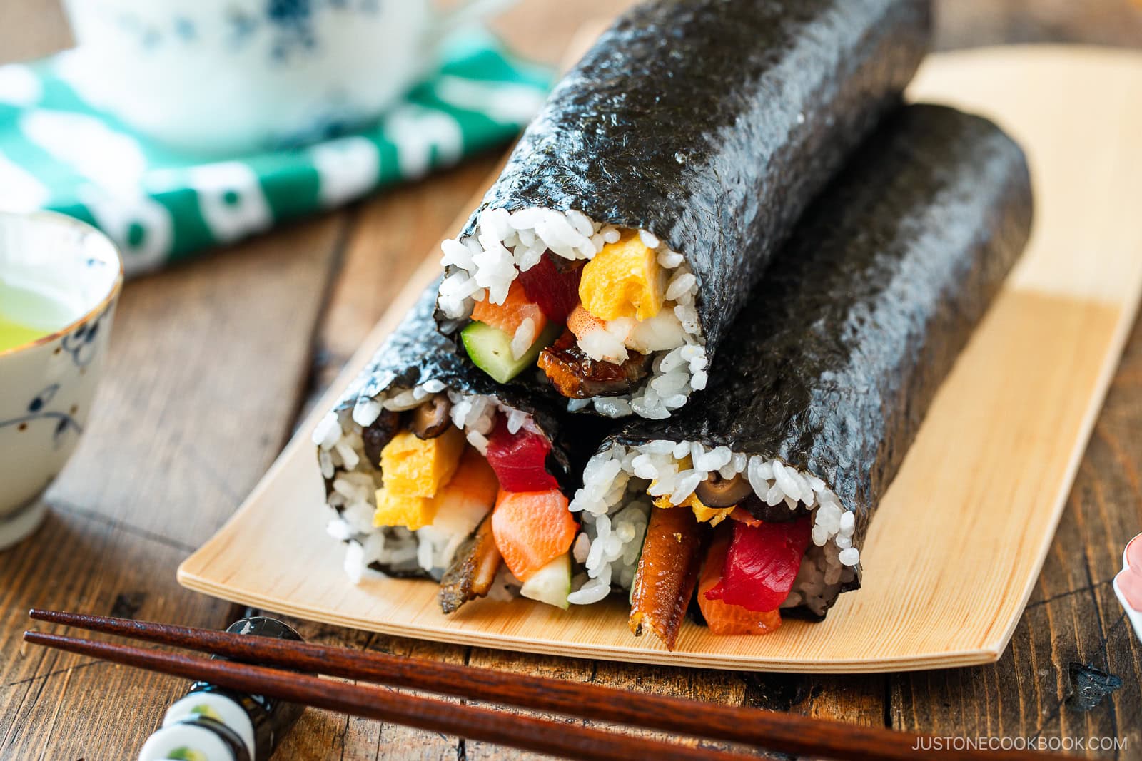 Two uncut sushi rolls—ehomaki (setsubun sushi roll)—filled with rice, vegetables, and seafood are wrapped in nori and arranged on a bamboo plate with chopsticks and a small teacup nearby.
