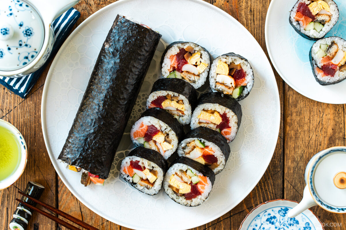 A white plate holds a whole ehomaki (setsubun sushi roll) and sliced pieces filled with rice, egg, vegetables, and fish. Surrounding the plate are small dishes, a teapot, a cup of green tea, and chopsticks on a wooden table.
