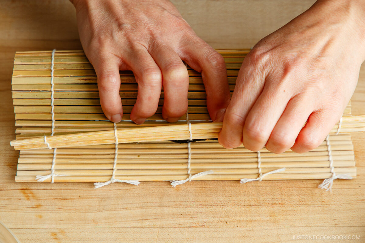 Close-up of hands using a bamboo sushi rolling mat to roll ehomaki (setsubun sushi roll) on a wooden surface. The mat is tied with string to hold it together.