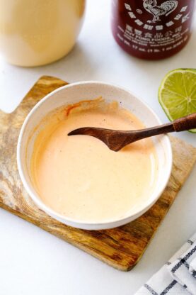 A bowl of creamy orange sauce, made from a homemade spicy mayo recipe, sits with a wooden spoon on a small board. Sriracha and mayonnaise bottles and a lime wedge are visible in the background.