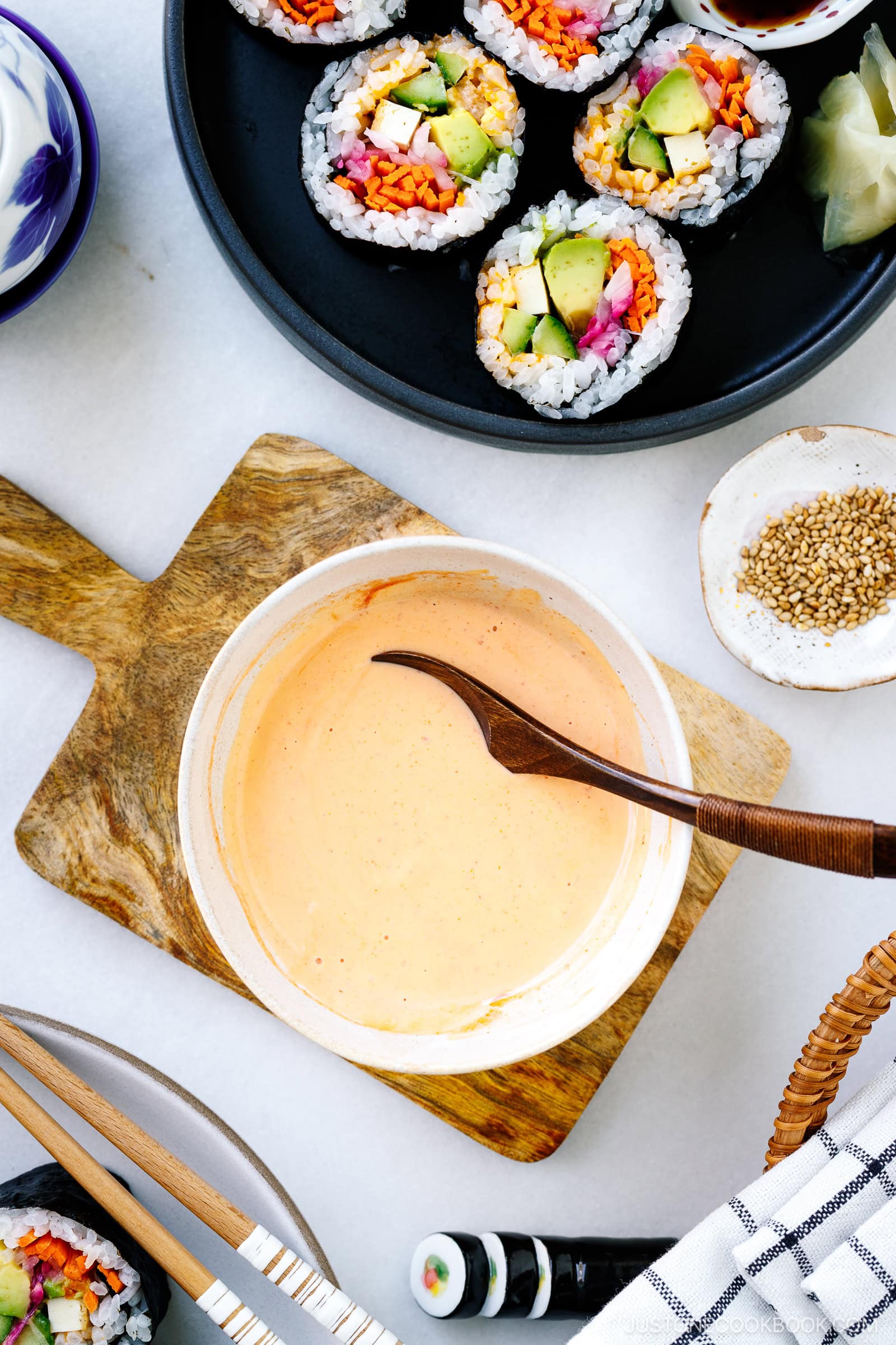 A bowl of creamy orange homemade spicy mayo recipe with a spoon sits on a wooden board, surrounded by sushi rolls filled with vegetables, sesame seeds in a small dish, and chopsticks on a white surface.