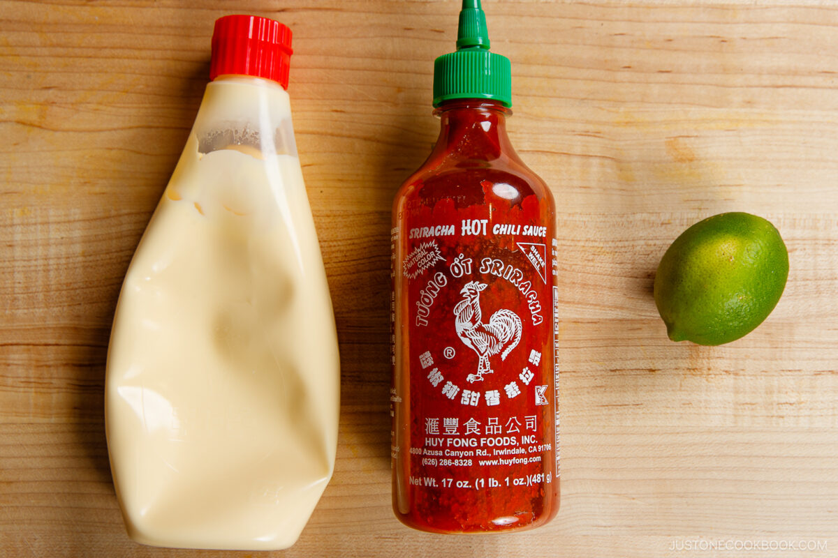 A bottle of mayonnaise, a bottle of Sriracha hot chili sauce, and a whole lime sit on a wooden surface—perfect ingredients for a homemade spicy mayo recipe.