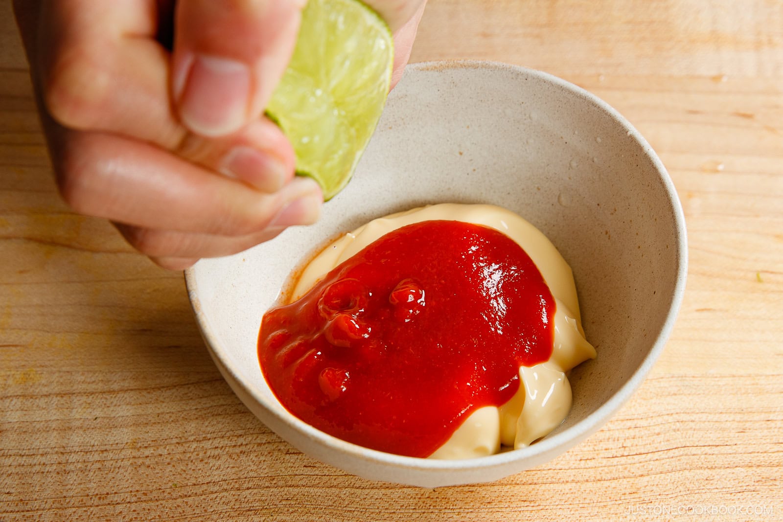 A hand squeezes a lime over a bowl containing mayonnaise and a generous amount of ketchup, crafting the perfect base for a homemade spicy mayo recipe on a wooden surface.