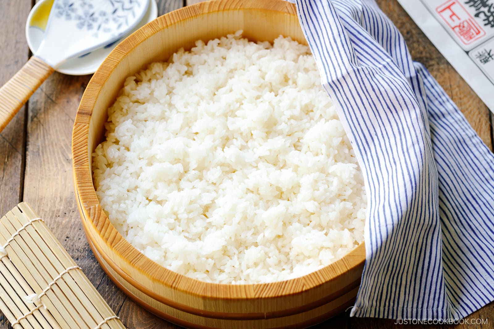A wooden bowl filled with cooked white sushi rice, perfect for learning how to make sushi rice, sits on a wooden table, partially covered by a blue and white striped cloth with a bamboo sushi mat and ceramic spoon nearby.