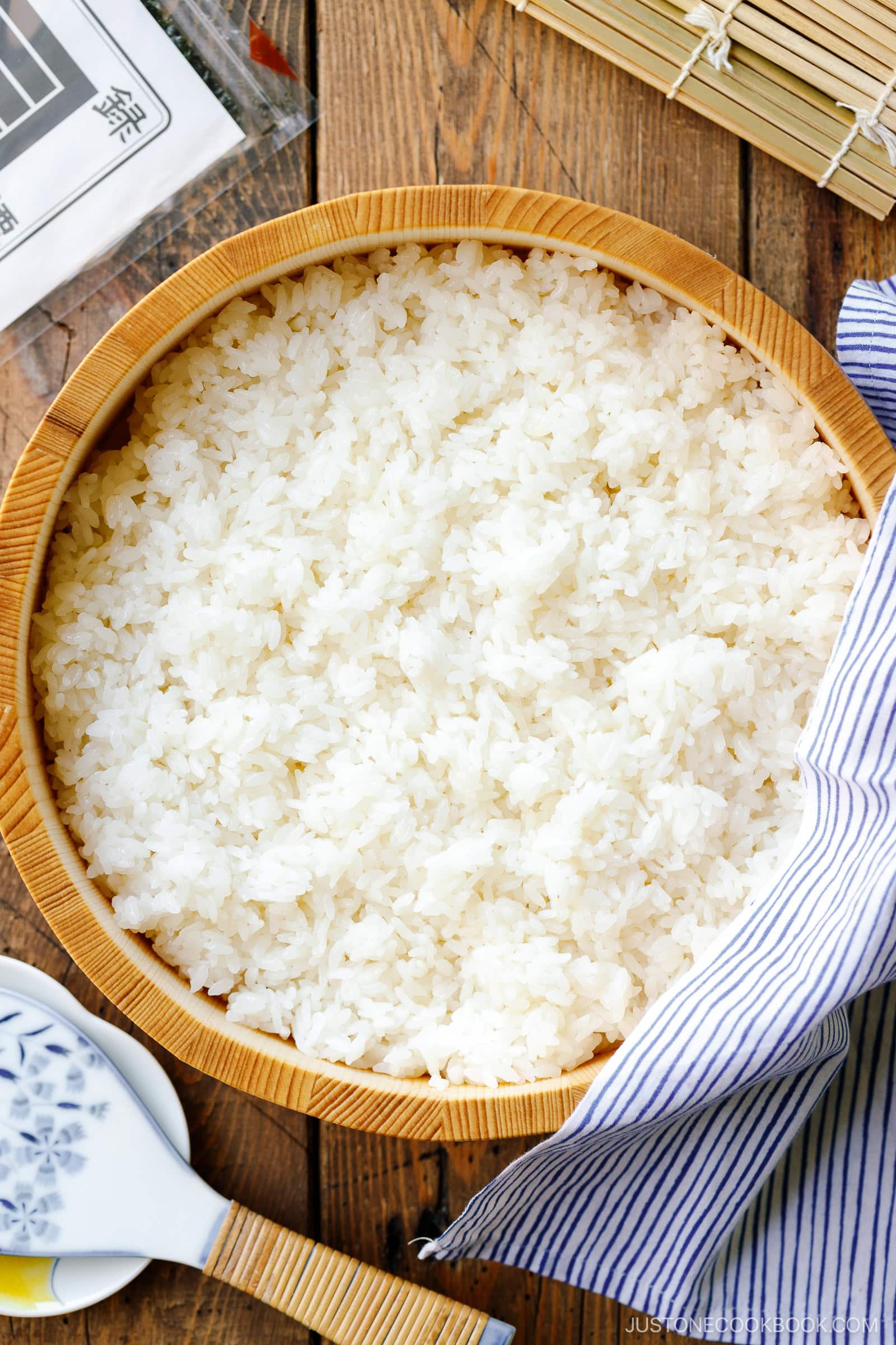 A large wooden bowl filled with cooked white rice sits on a wooden table, partially covered by a blue-and-white striped cloth—perfect for anyone learning how to make sushi rice. Nearby are a rice paddle and a sushi rolling mat.