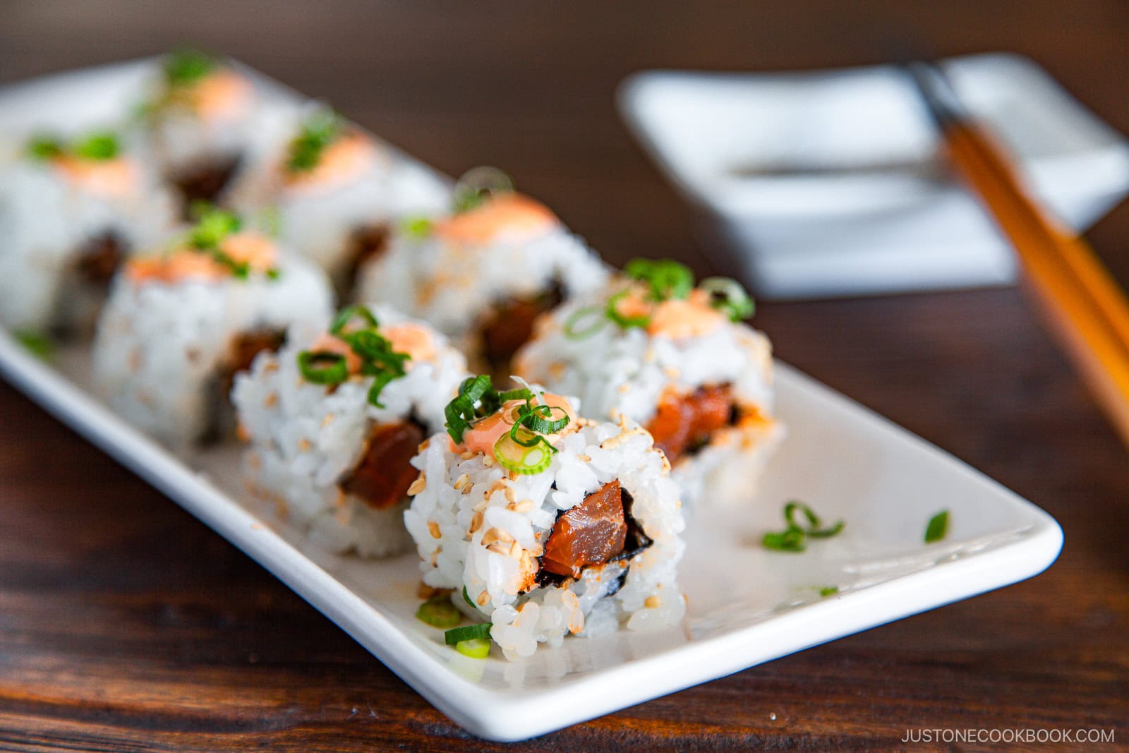 Close-up of a rectangular white plate featuring spicy tuna rolls topped with chopped green onions and spicy mayo, filled with fish and rice. Chopsticks and a small dish are in the background.