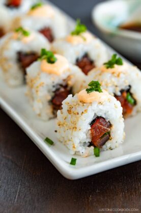Close-up of spicy tuna rolls topped with creamy orange sauce and chopped green onions, arranged neatly on a white rectangular plate.