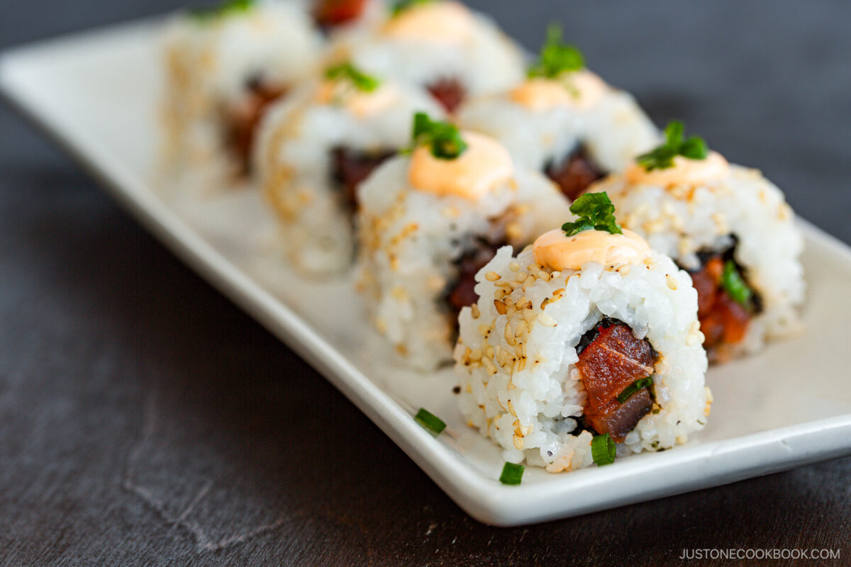 A rectangular white plate holds a row of spicy tuna rolls, each topped with a dollop of creamy orange sauce and sprinkled with chopped green onions and sesame seeds. The background is a dark, blurred surface.
