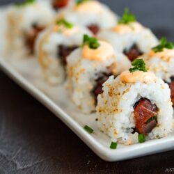 A rectangular white plate holds a row of spicy tuna rolls, each topped with a dollop of creamy orange sauce and sprinkled with chopped green onions and sesame seeds. The background is a dark, blurred surface.