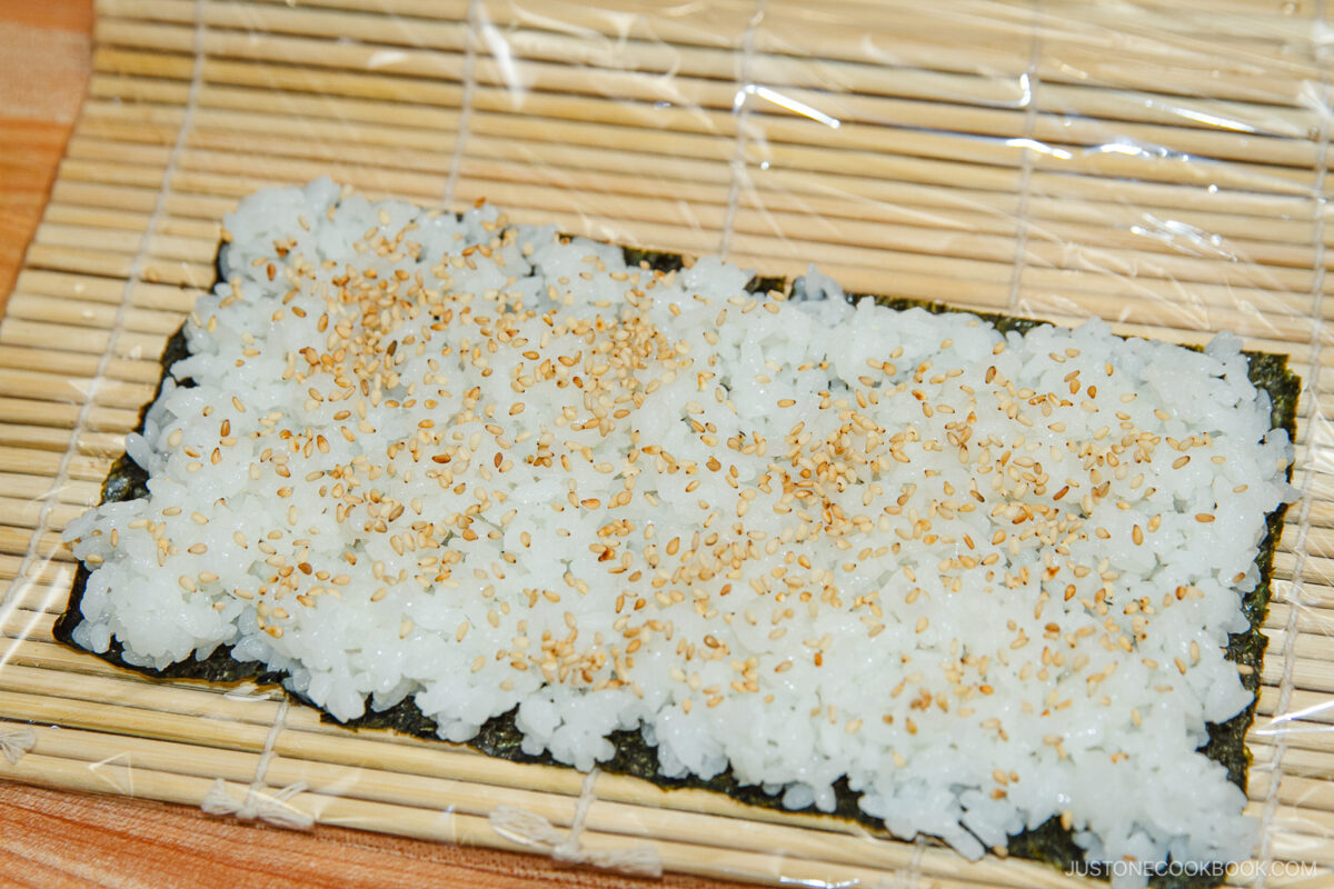 A bamboo sushi rolling mat with a sheet of nori covered in a layer of white sushi rice, sprinkled with sesame seeds, ready to craft delicious spicy tuna rolls.