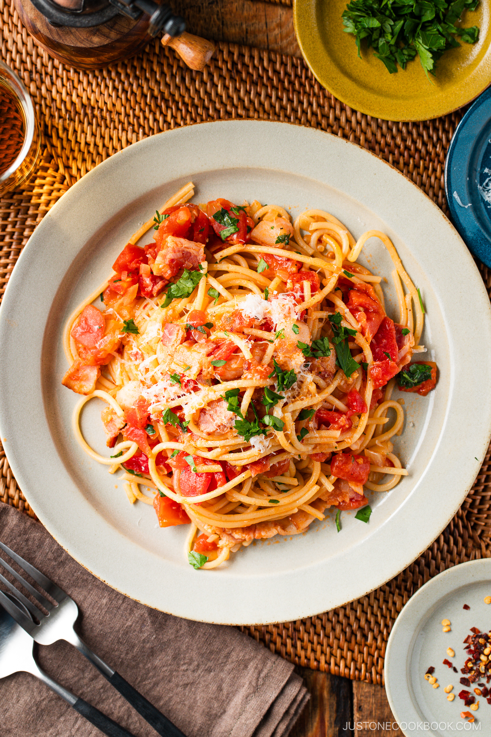 A plate of Tomato Bacon Pasta topped with diced tomatoes, grated cheese, and fresh parsley sits on a woven placemat, surrounded by cutlery, a napkin, chopped herbs, and a pepper grinder.