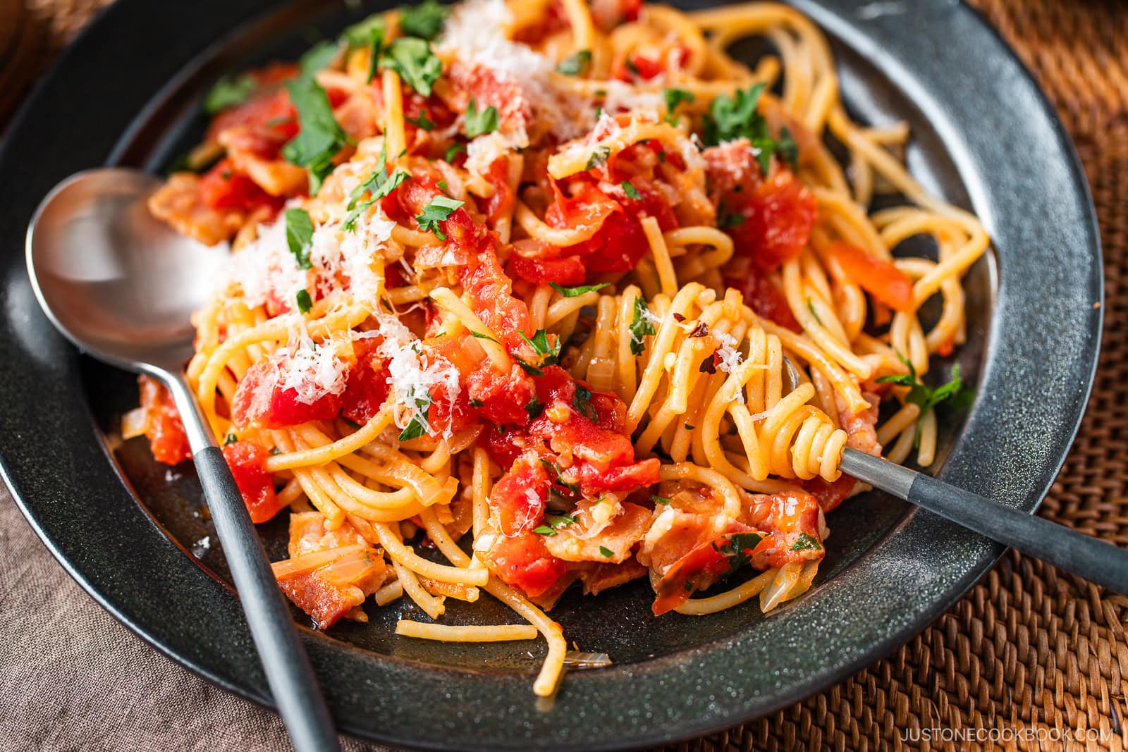 A plate of Tomato Bacon Pasta topped with tomato sauce, chopped tomatoes, herbs, and grated cheese, with a fork and spoon resting on the side. The dish is served on a dark plate with a woven placemat underneath.