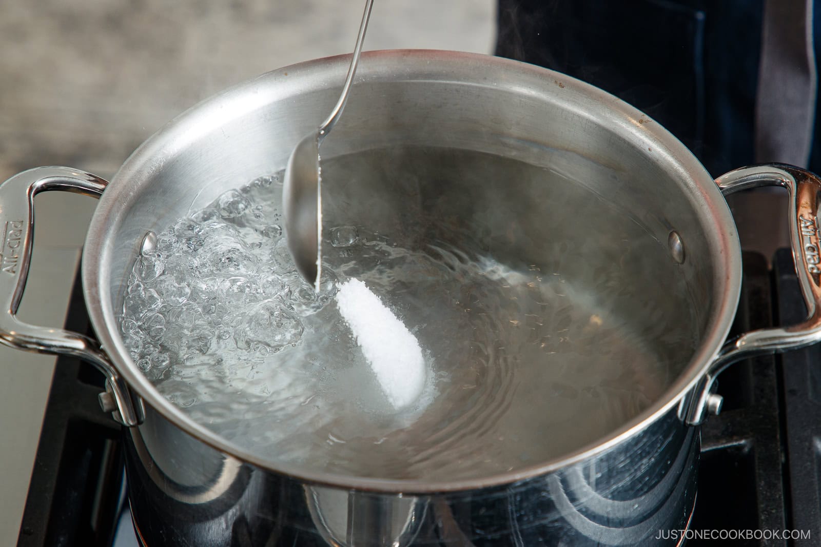 A spoonful of salt is being added to a pot of boiling water on the stove, preparing the base for a delicious Tomato Bacon Pasta. Steam rises as the salt is about to dissolve.