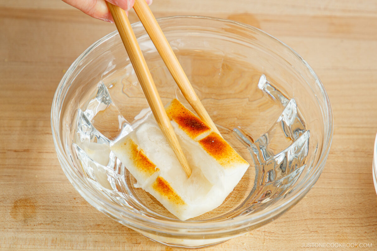 A hand uses wooden chopsticks to hold a piece of grilled kinako mochi in a clear glass bowl on a wooden surface.