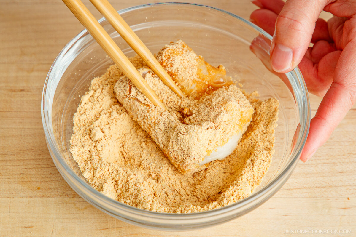 A hand uses chopsticks to coat a piece of kinako mochi with light brown soybean flour in a clear glass bowl on a wooden surface.