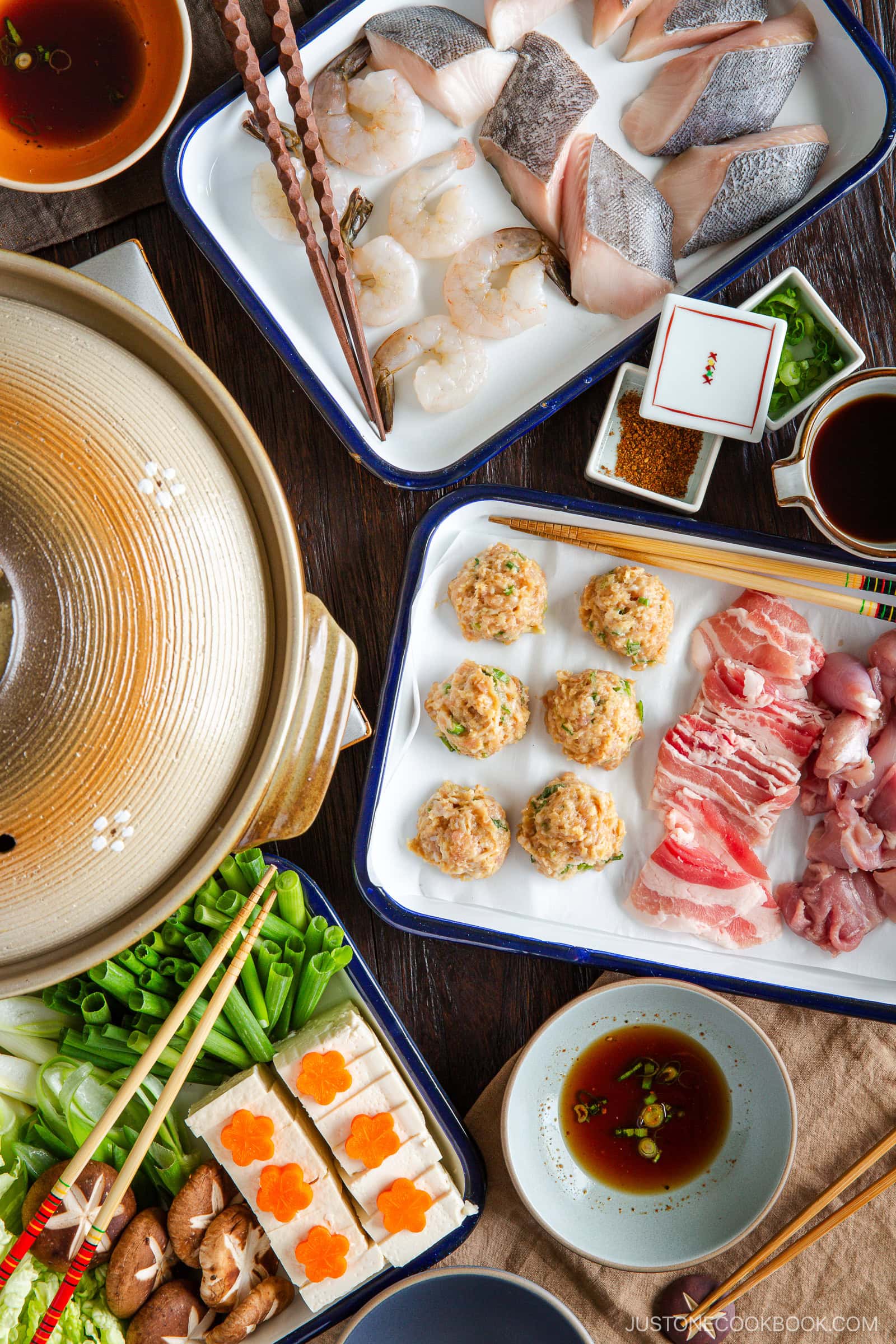 A Japanese hot pot meal featuring chanko nabe (sumo stew) with plates of raw seafood, meat, meatballs, tofu with carrot slices, mushrooms, green onions, dipping sauces, and a pot of broth ready for cooking.