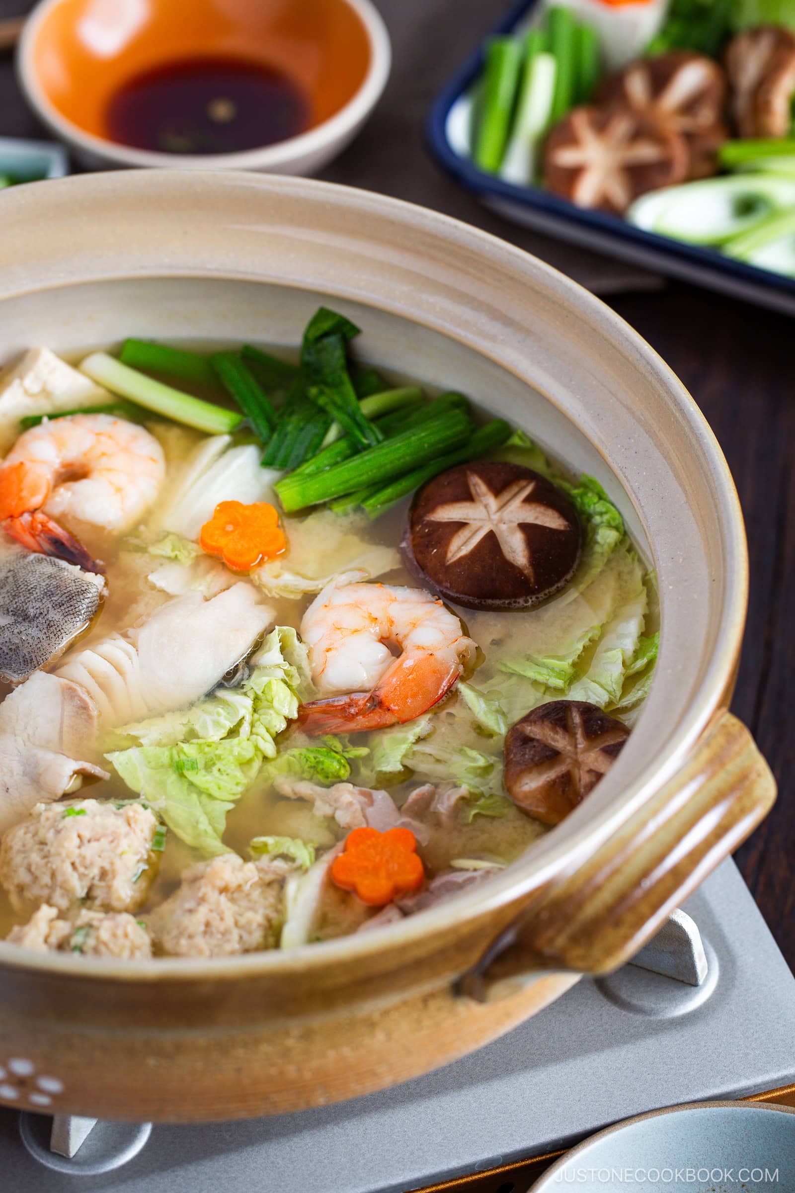 A hot pot of chanko nabe (sumo stew) filled with shrimp, fish, meatballs, mushrooms, cabbage, green onions, and carrot slices sits on a portable stove, with dipping sauce and more vegetables on plates in the background.