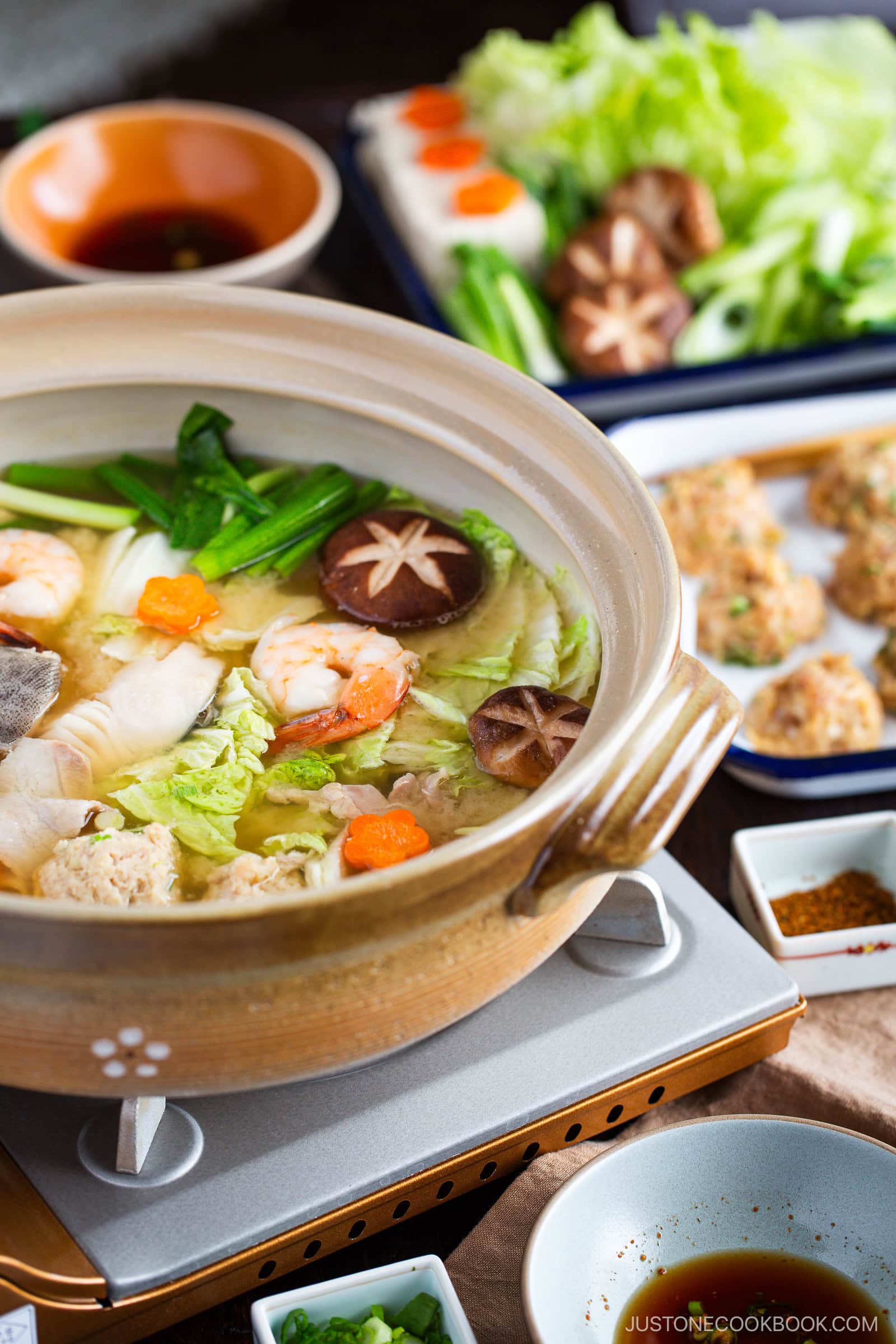 A simmering clay pot of Japanese chanko nabe (sumo stew) brims with vegetables, shrimp, mushrooms, and fish on a portable stove. Surrounding dishes hold fresh greens, dipping sauces, and meatballs ready to be cooked.