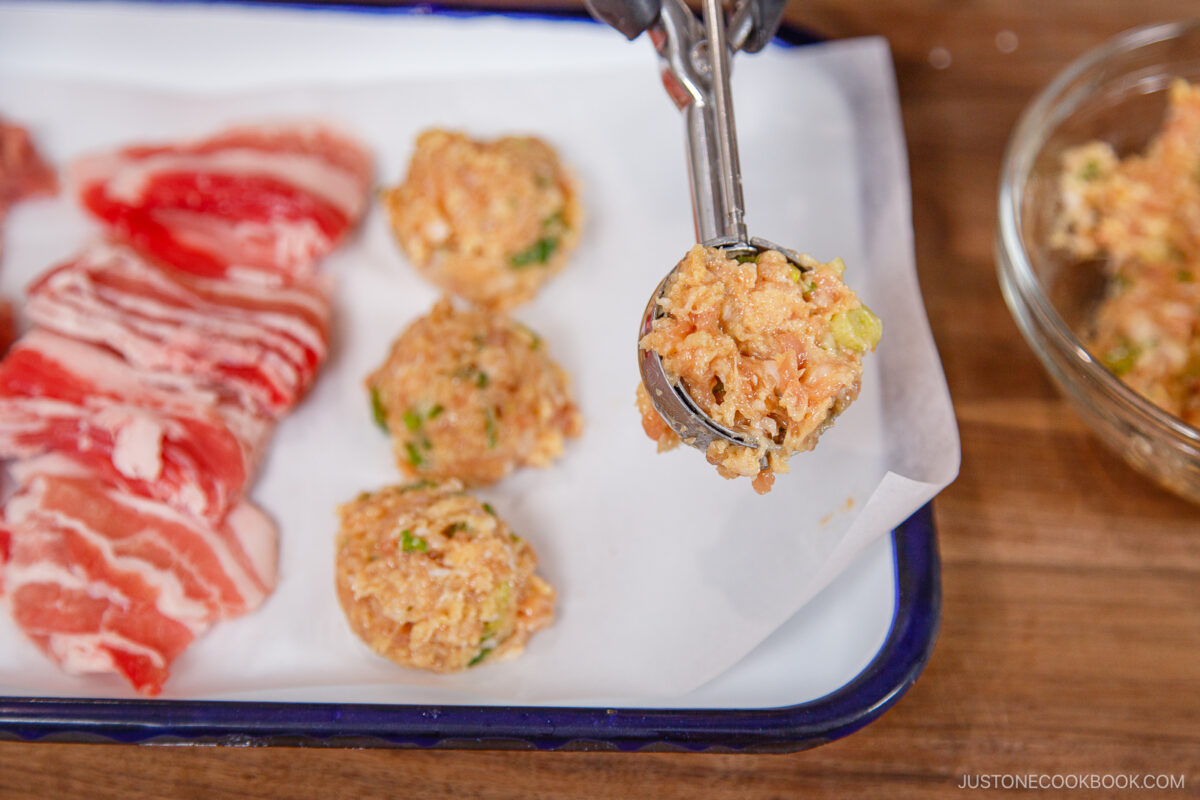 A hand uses a scoop to form meatballs for chanko nabe (sumo stew) from a bowl of mixture, placing them on a parchment-lined tray next to slices of raw beef.