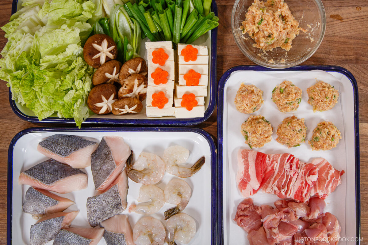 Various hot pot ingredients are arranged on trays, including leafy greens, shiitake mushrooms, tofu with carrot slices, fish fillets, shrimp, meatballs, sliced beef, and chicken pieces—perfect for making a hearty chanko nabe (sumo stew). A bowl with mixed filling is nearby.