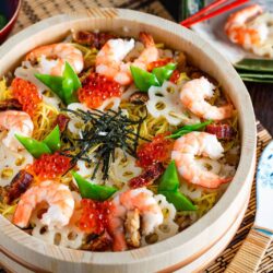 A round wooden dish of chirashi sushi is topped with shrimp, sliced lotus root, snap peas, eel, shredded nori, and bright orange salmon roe. A patterned rice paddle and a plate with chopsticks are placed beside the vibrant chirashi sushi.