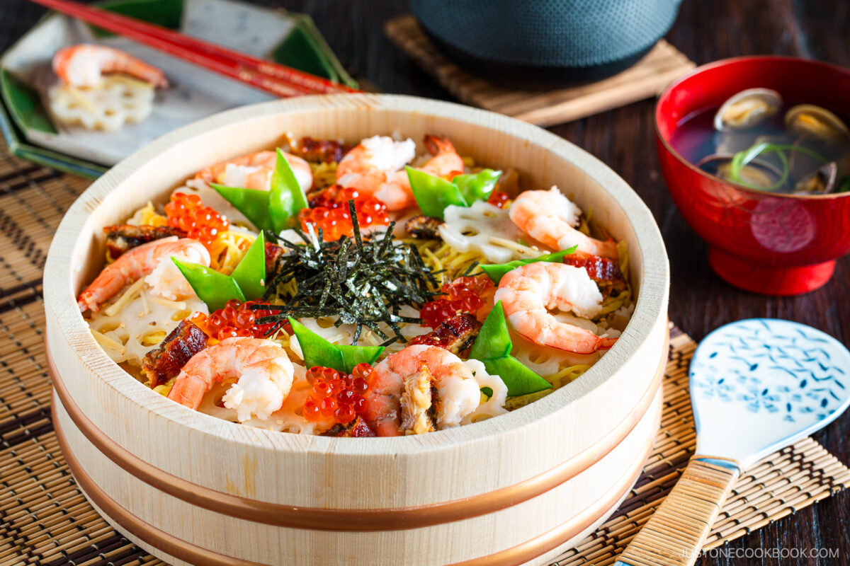 A wooden bowl of colorful chirashi sushi topped with shrimp, eel, lotus root, snow peas, salmon roe, and shredded seaweed is served with a bowl of soup, red chopsticks, and a rice paddle on a bamboo mat.