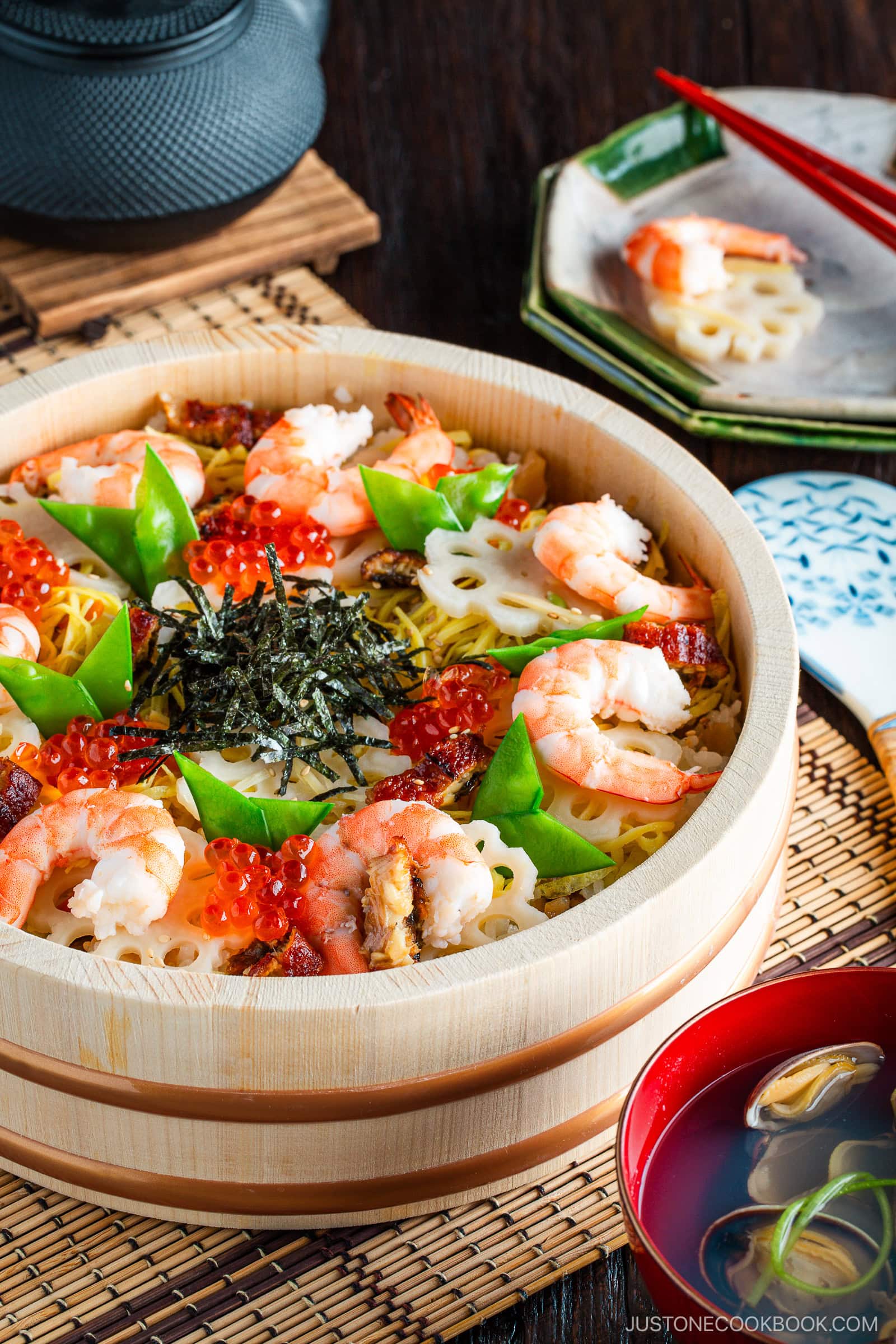 A wooden sushi oke of chirashi sushi features colorful shrimp, ikura (salmon roe), sliced lotus root, snow peas, egg, and seaweed, served with clear soup, chopsticks, and a teapot on a bamboo mat.