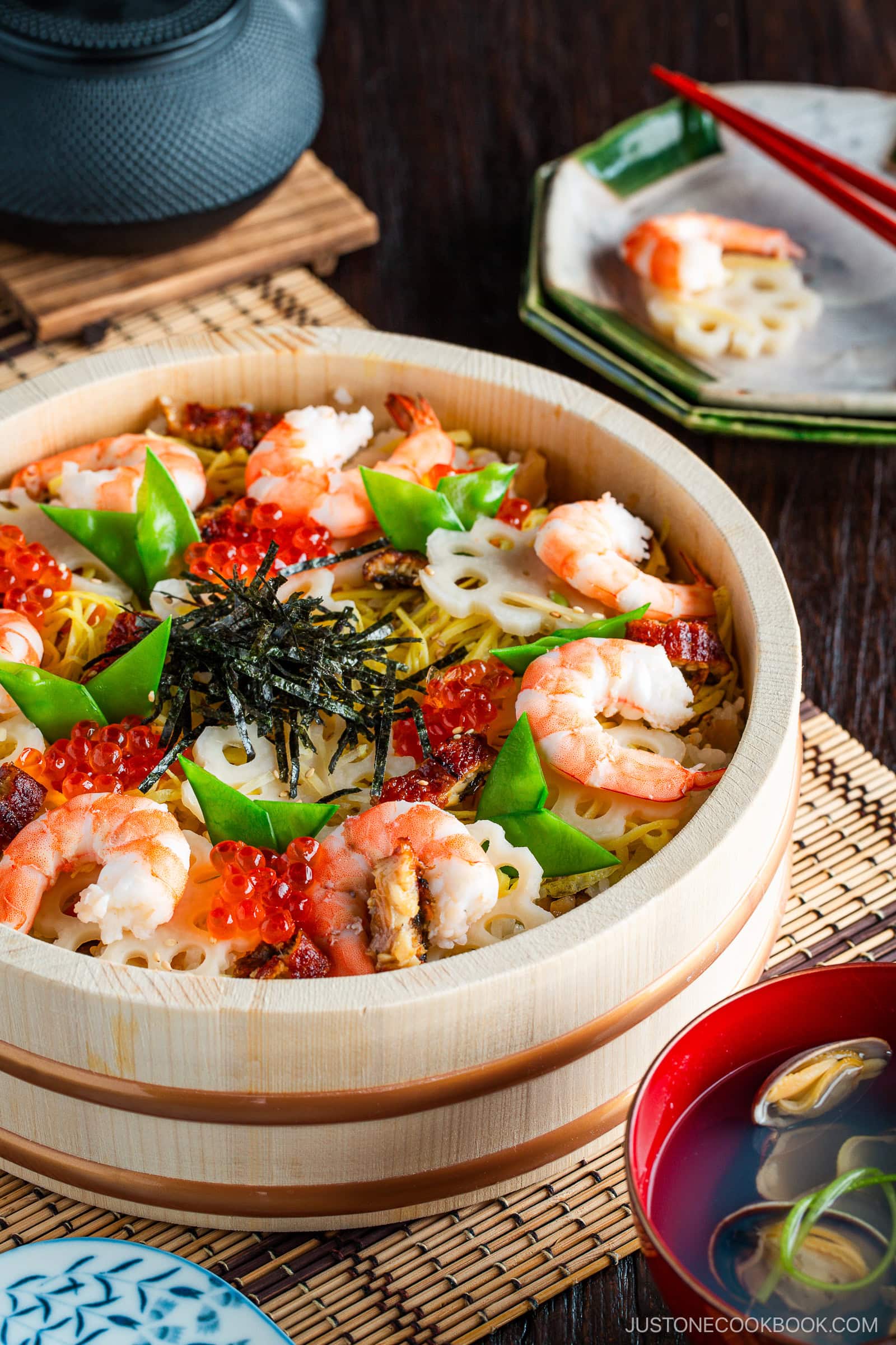 A wooden bowl filled with vibrant chirashi sushi—topped with shrimp, lotus root slices, snow peas, salmon roe, nori strips, and grilled eel—served alongside miso soup and a small plate with chopsticks in the background.