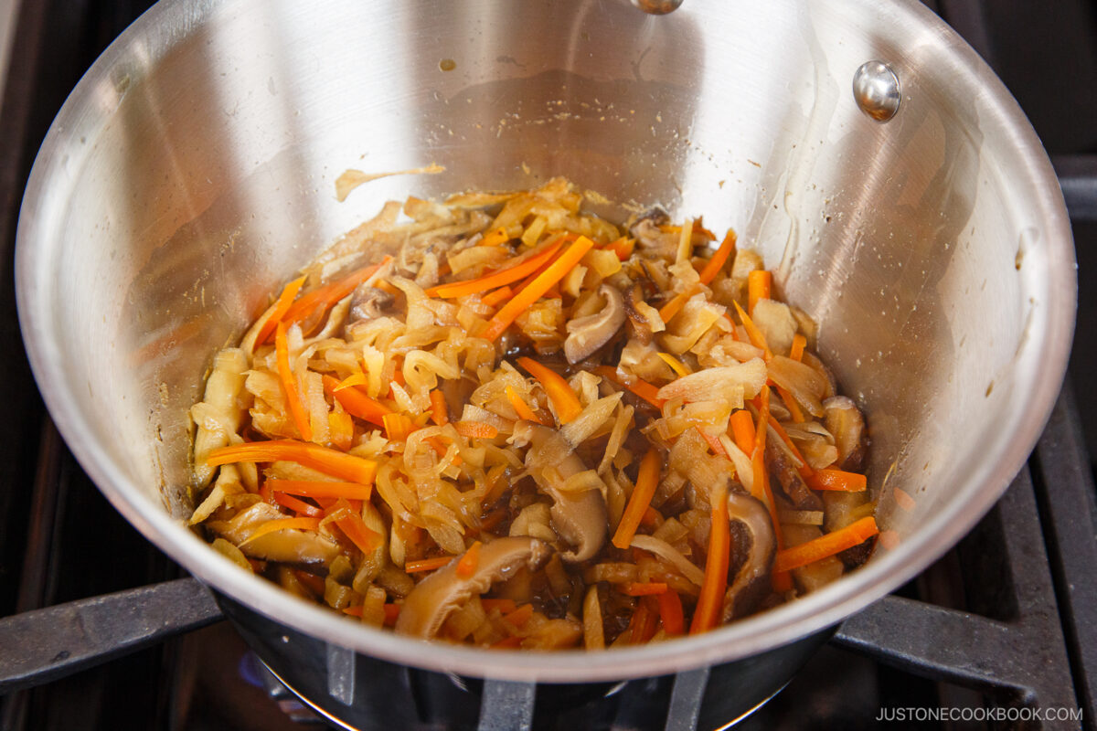 A stainless steel pot on a stove contains sautéed onions, carrots, and mushrooms, lightly caramelized and mixed together—perfect vegetables to add a savory depth to your homemade chirashi sushi.
