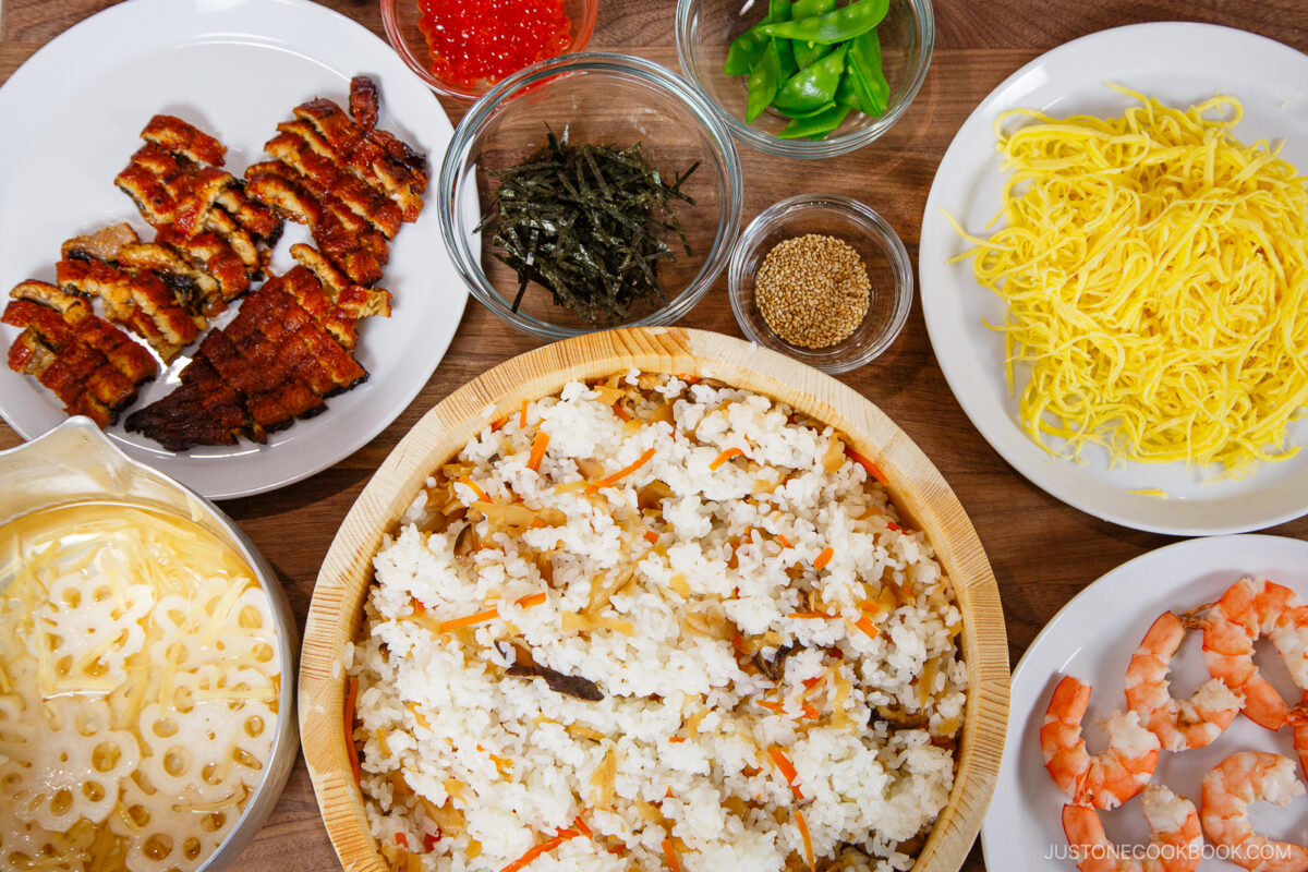 A top-down view of assorted bowl and plate ingredients for making chirashi sushi, including rice, eel, shrimp, egg strips, lotus root, seaweed strips, snap peas, and fish roe.