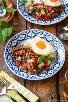 A plate of gapao rice features stir-fried ground meat with onions, red bell peppers, herbs, and a sunny-side-up egg on top, served over rice in a blue and white patterned bowl on a wooden table.