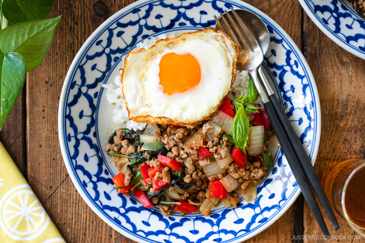 A blue and white patterned bowl filled with gapao rice—stir-fried ground meat, onions, red bell peppers, basil, and rice—topped with a sunny-side-up egg. A fork and spoon rest on the side of the bowl.