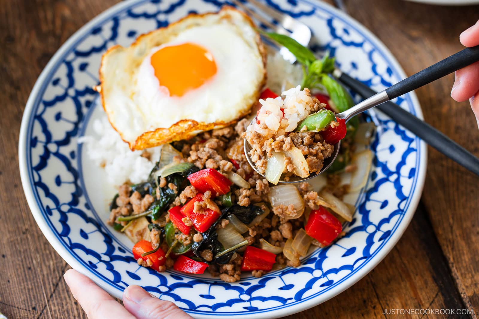 A plate of gapao rice features stir-fried minced meat with red bell peppers, onions, and basil over white rice, topped with a sunny-side-up egg. A hand holds a spoonful of the dish above the patterned blue and white plate.