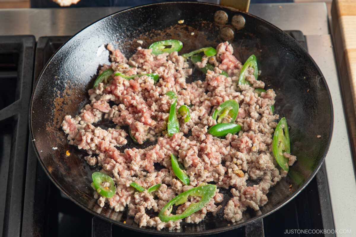 Ground meat and sliced green peppers are being cooked in a black frying pan on a stovetop, similar to the preparation of gapao rice. The meat is browning as the peppers are scattered throughout the pan.