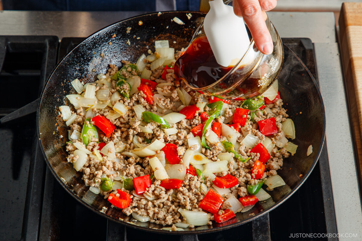 A hand pours dark sauce from a small pitcher into a skillet with cooked ground meat, chopped onions, green bell peppers, and red bell peppers on a stovetop—perfect flavors for making gapao rice.