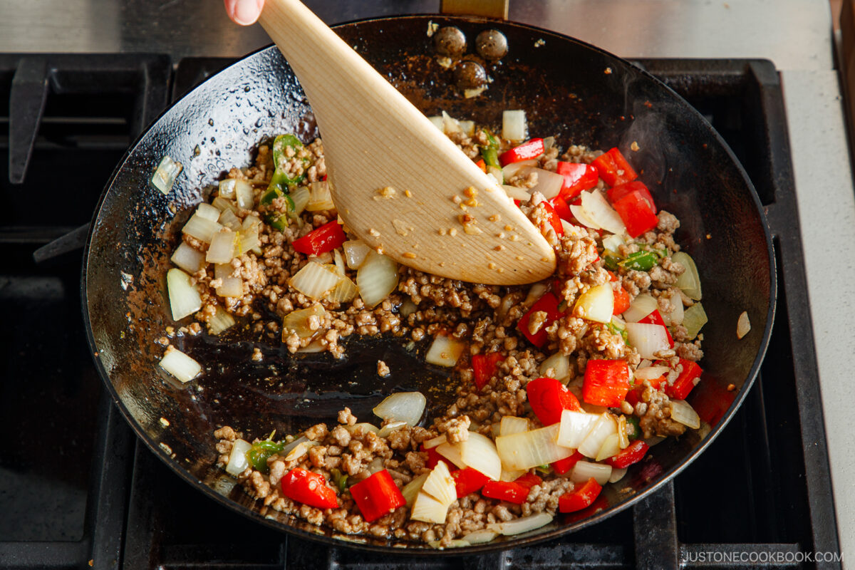 A wooden spatula stirs ground meat, chopped onions, and red bell peppers in a black skillet on a stovetop, creating the flavorful base for gapao rice.