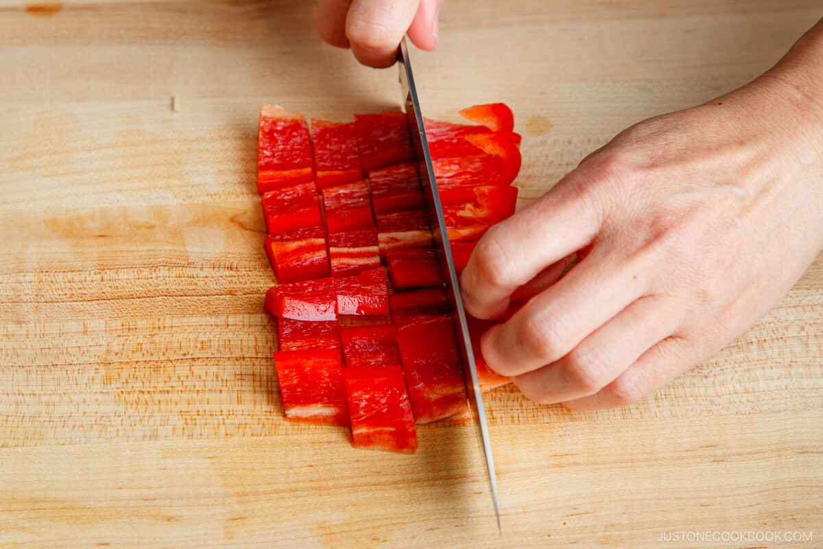 A person slices red bell pepper into small square pieces on a wooden cutting board with a sharp knife, preparing fresh ingredients for gapao rice.
