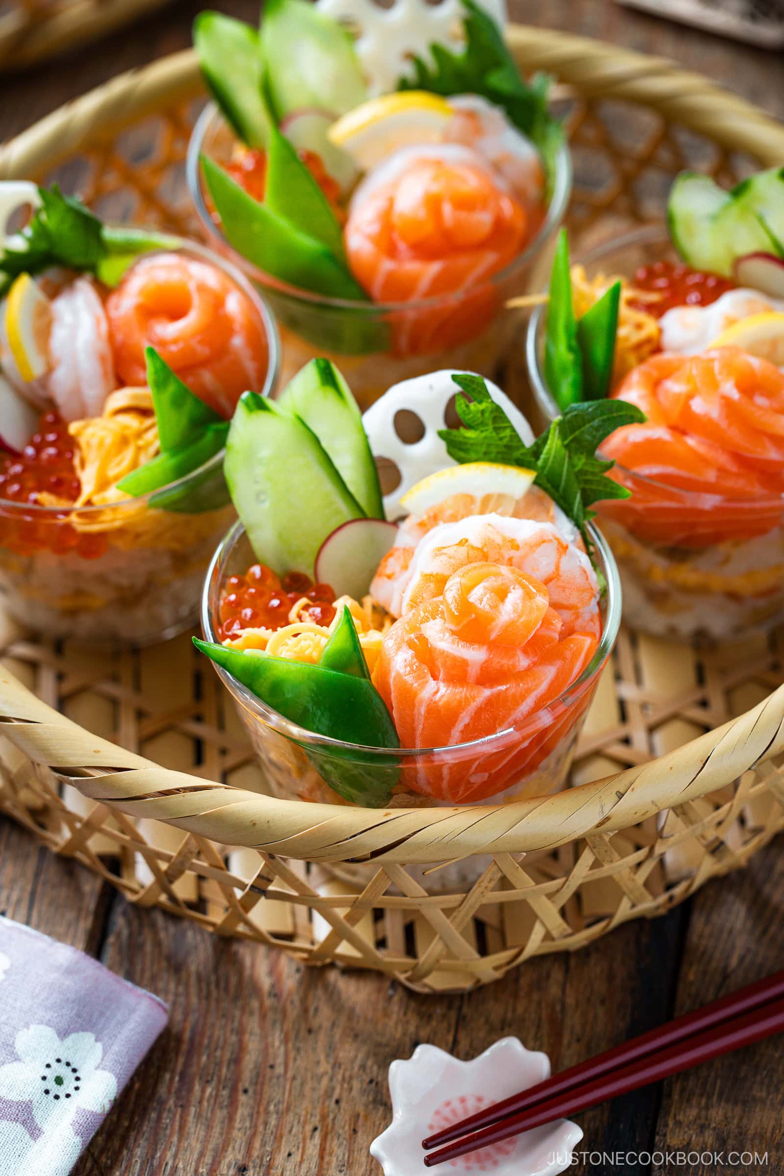 Quick & Easy Chirashi Sushi cups filled with layers of sushi rice, salmon roses, shrimp, shredded egg, cucumber, lotus root slices, and ikura are arranged in a woven bamboo tray on a wooden table with chopsticks nearby.