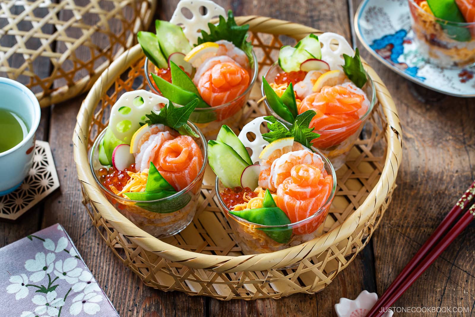 Six clear cups filled with colorful Quick & Easy Chirashi Sushi ingredients like salmon, shrimp, cucumber, ikura, egg, radish, and lotus root are arranged neatly in a woven bamboo basket on a wooden table with tea and chopsticks nearby.