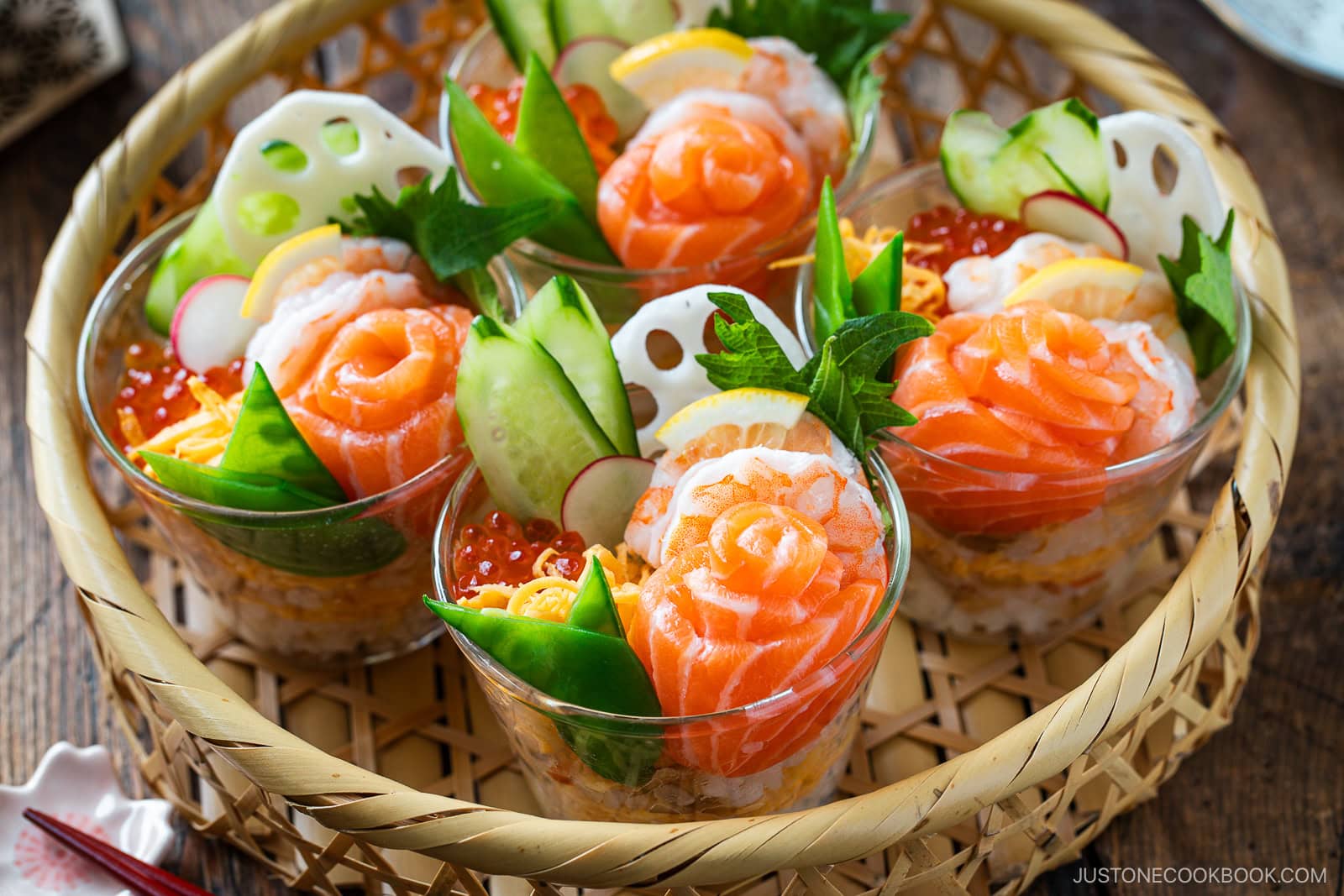 Individual glass cups showcase Quick & Easy Chirashi Sushi, topped with salmon, shrimp, cucumber, radish, snap peas, lotus root, salmon roe, lemon slices, and fresh green garnish—all artfully arranged in a woven basket.