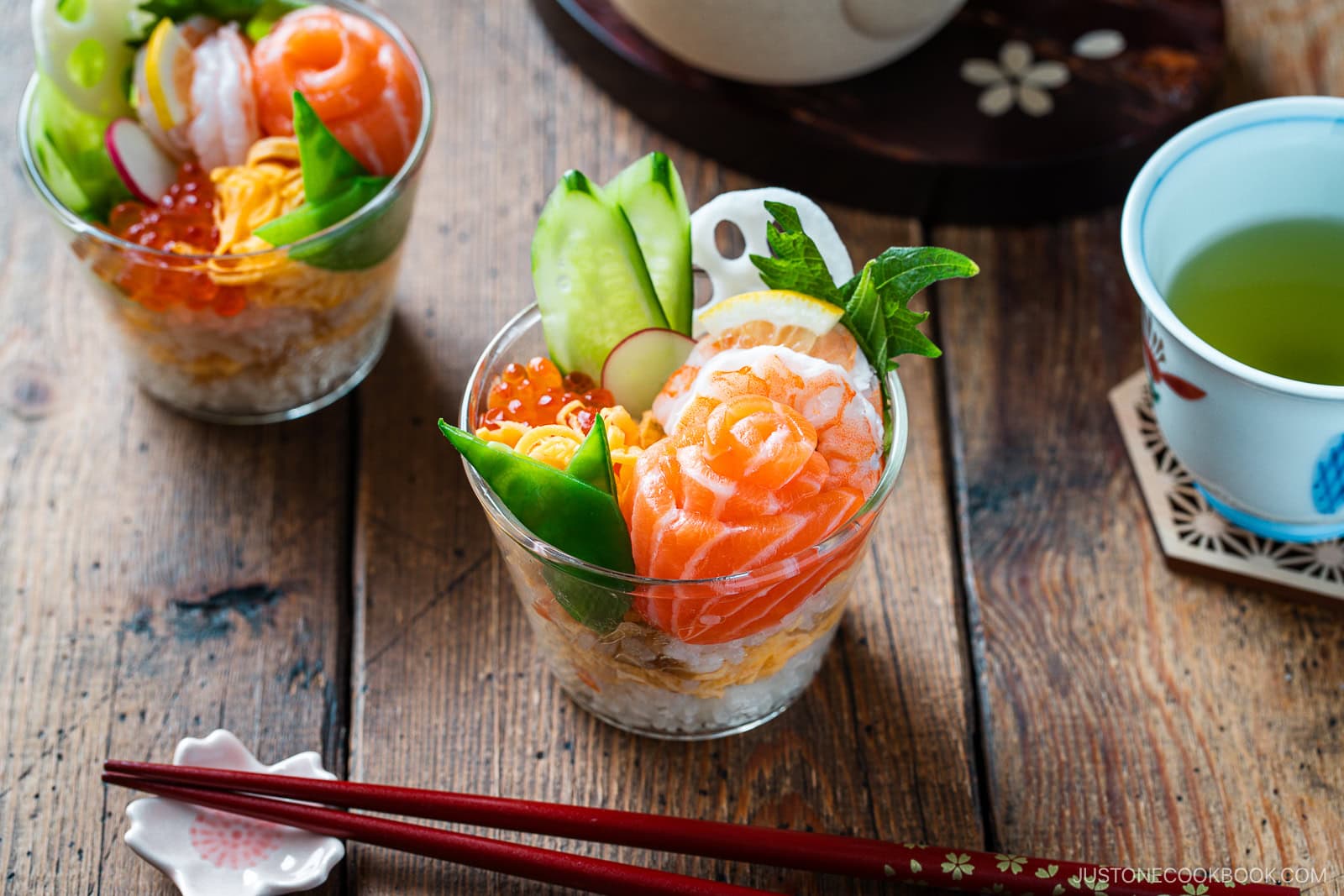 A glass cup filled with Quick & Easy Chirashi Sushi—sushi rice, sliced salmon, shrimp, egg, cucumber, snap peas, salmon roe, and lotus root—garnished with greens on a wooden table beside red chopsticks and green tea.