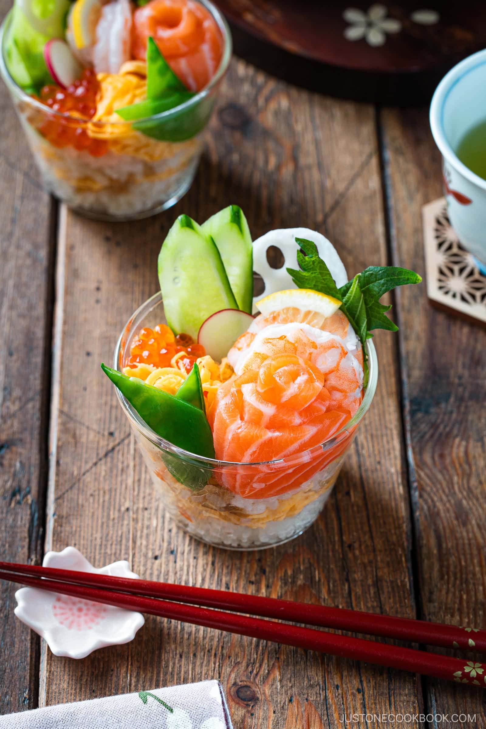 A glass cup filled with sushi rice, topped with salmon sashimi, cucumber slices, lotus root, ikura (salmon roe), and green leaves—this Quick & Easy Chirashi Sushi is served on a wooden table with red chopsticks beside it.